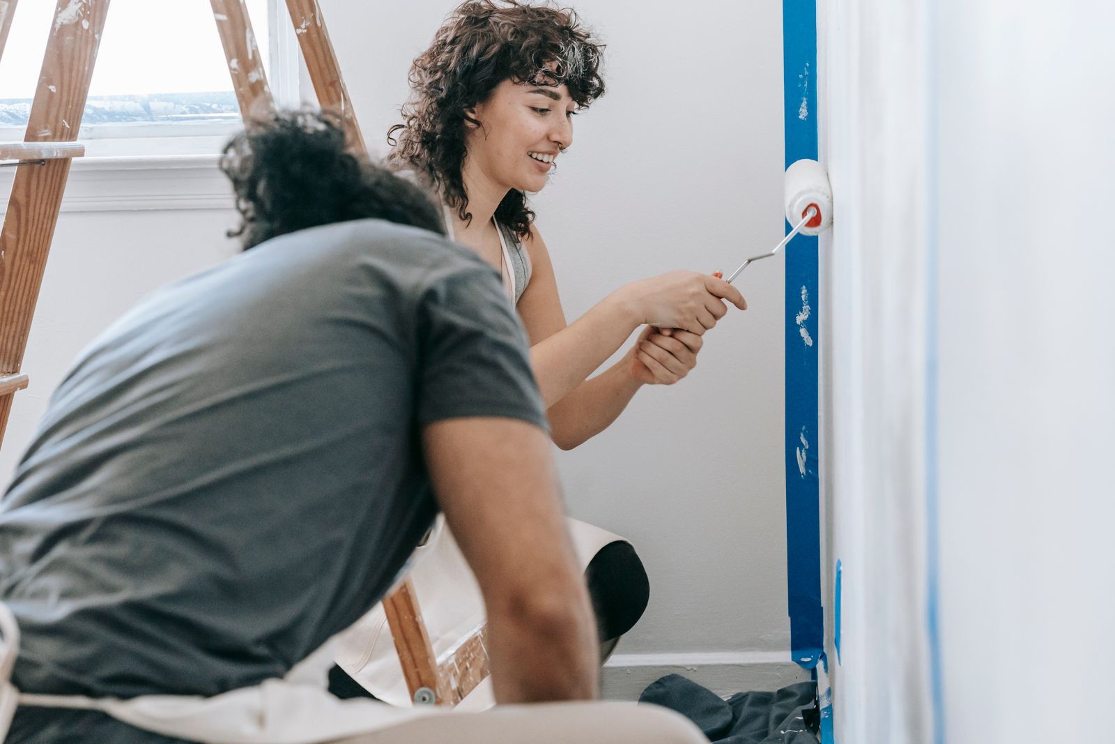 A two-person painting crew rolling a fresh coat onto the walls of a bright interior room, with painter's tape and drop cloths in place